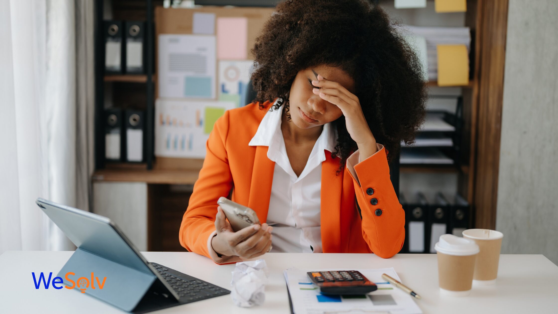 Black woman in an orange blazer with her hand on her forehead, looking at her phone very frustrated.